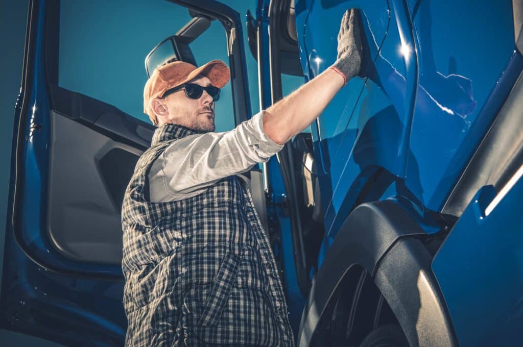 Male truck driver wearing hat and sunglasses, running gloved hand along side of truck cab