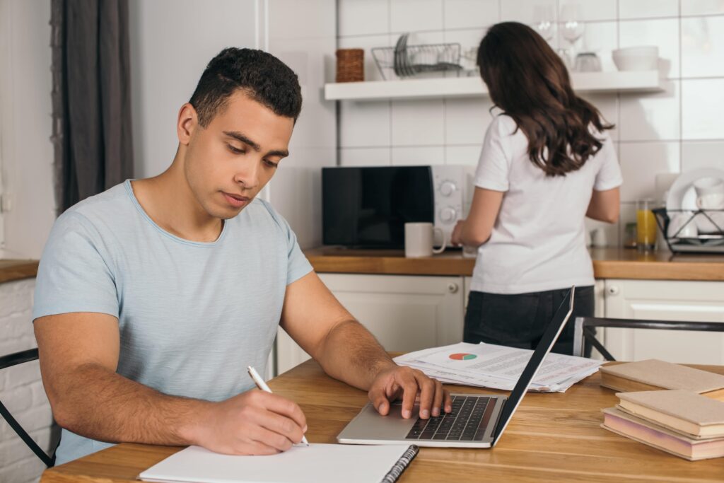 Man studying using laptop and notebook, wife in kitchen behind him